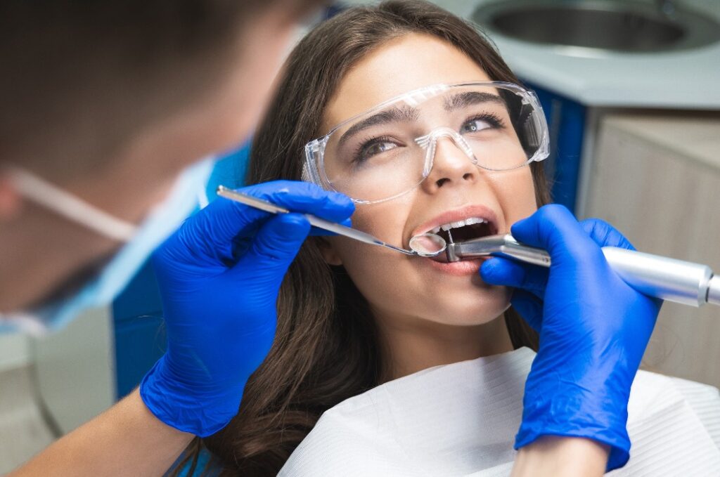 Zahnarzt mit Maske füllt den Wurzelkanal der Patientin, während sie mit Schutzbrille unter der medizinischen Lampe in der Klinik auf dem Zahnarztstuhl liegt